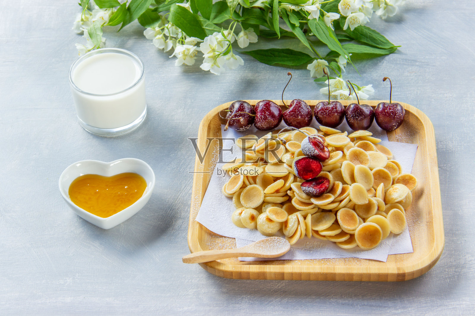 小煎饼麦片,樱桃,牛奶和蜂蜜。在明亮的背景上装饰着茉莉花的小枝。时髦的食物,家庭早餐。给孩子们的有趣食物。照片摄影图片