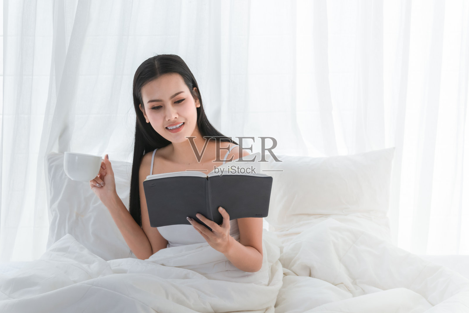 亚洲女孩可爱和美丽在床上看书和拿着咖啡杯在快乐,放松时间或工作为家。照片摄影图片