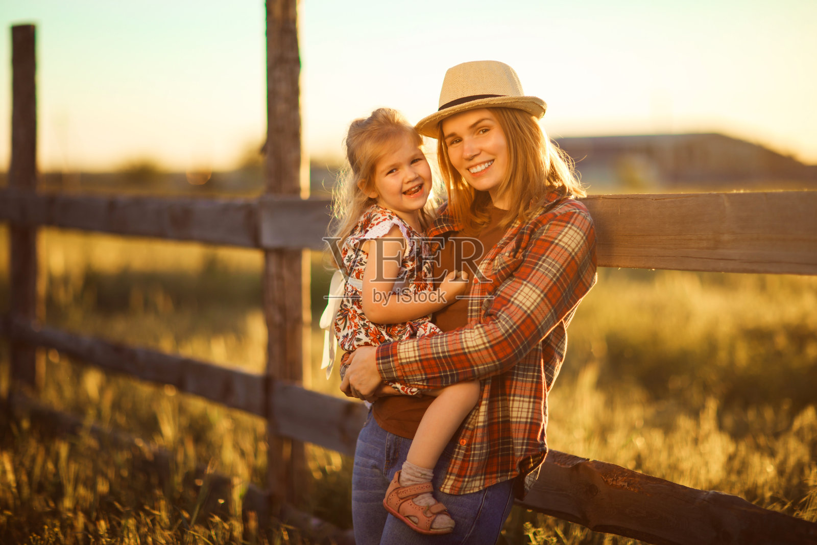 母亲和女儿戴着帽子,格子衬衫站在篱笆附近在日落的村庄照片摄影图片