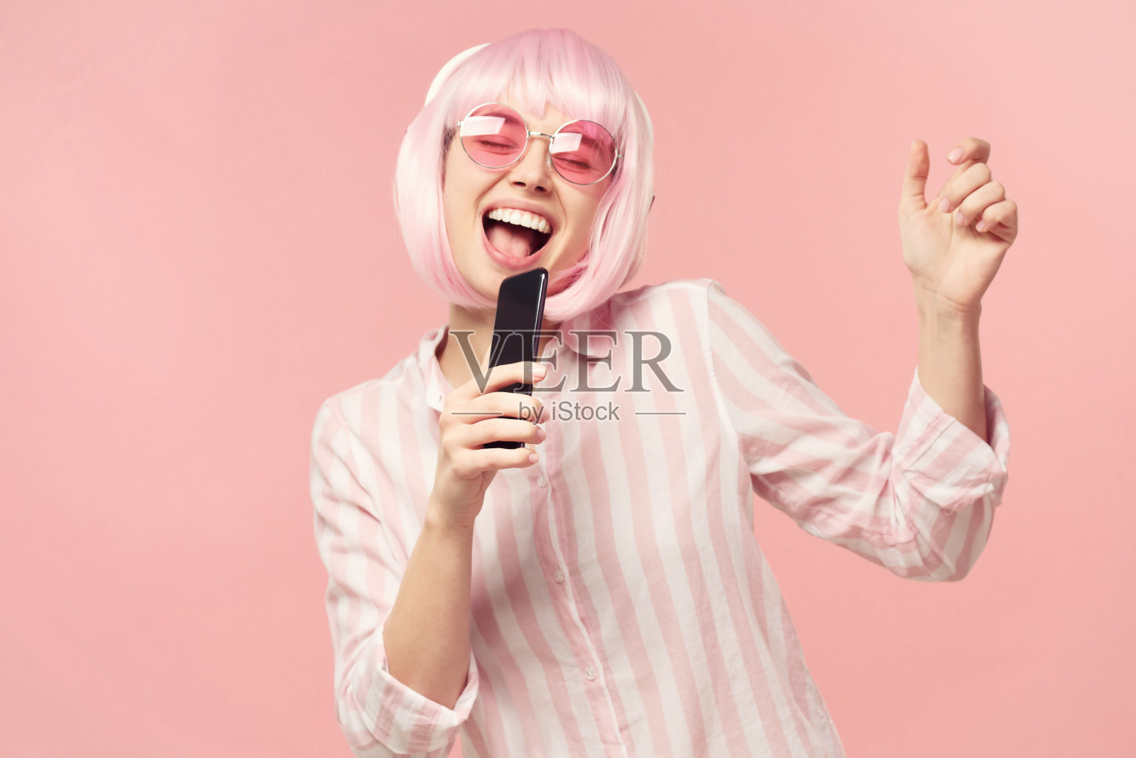 快乐的年轻女性拿着智能手机,在在线卡拉ok应用中唱歌,在粉红色的背景上孤立照片摄影图片