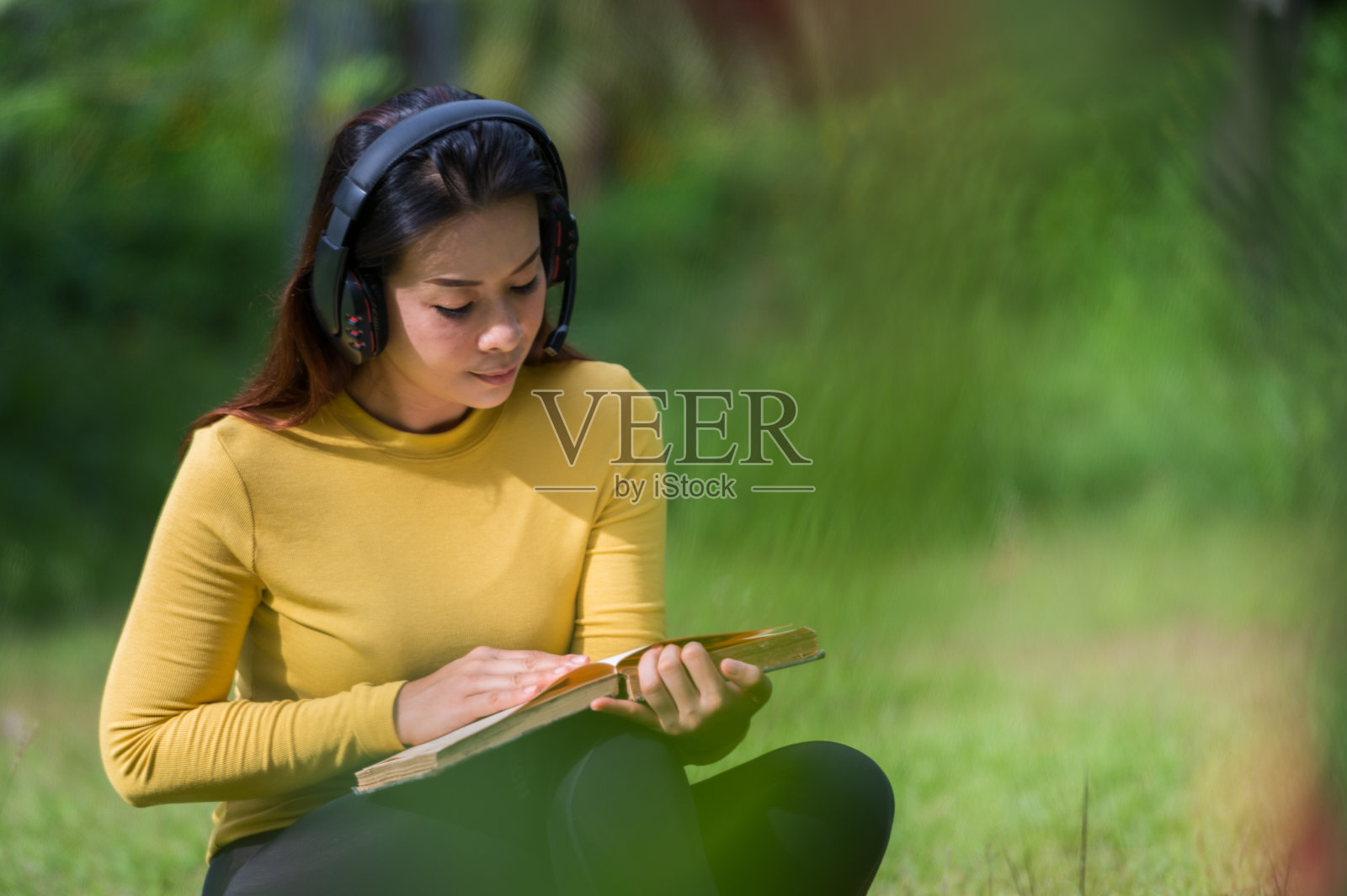 女学生穿着黄色的衬衫,拿着书,戴着耳机,坐在绿色的草坪上。照片摄影图片