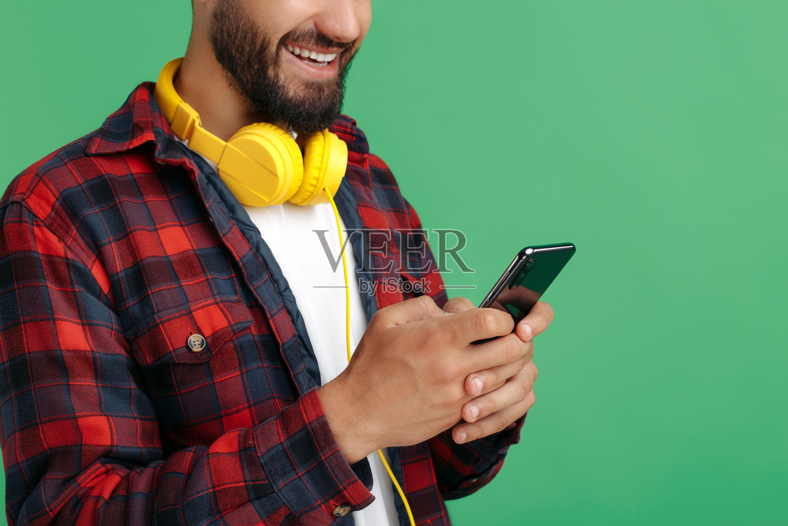 一名快乐的留着胡子的潮男穿着格子衬衫,戴着耳机在绿色背景上使用智能手机。照片摄影图片