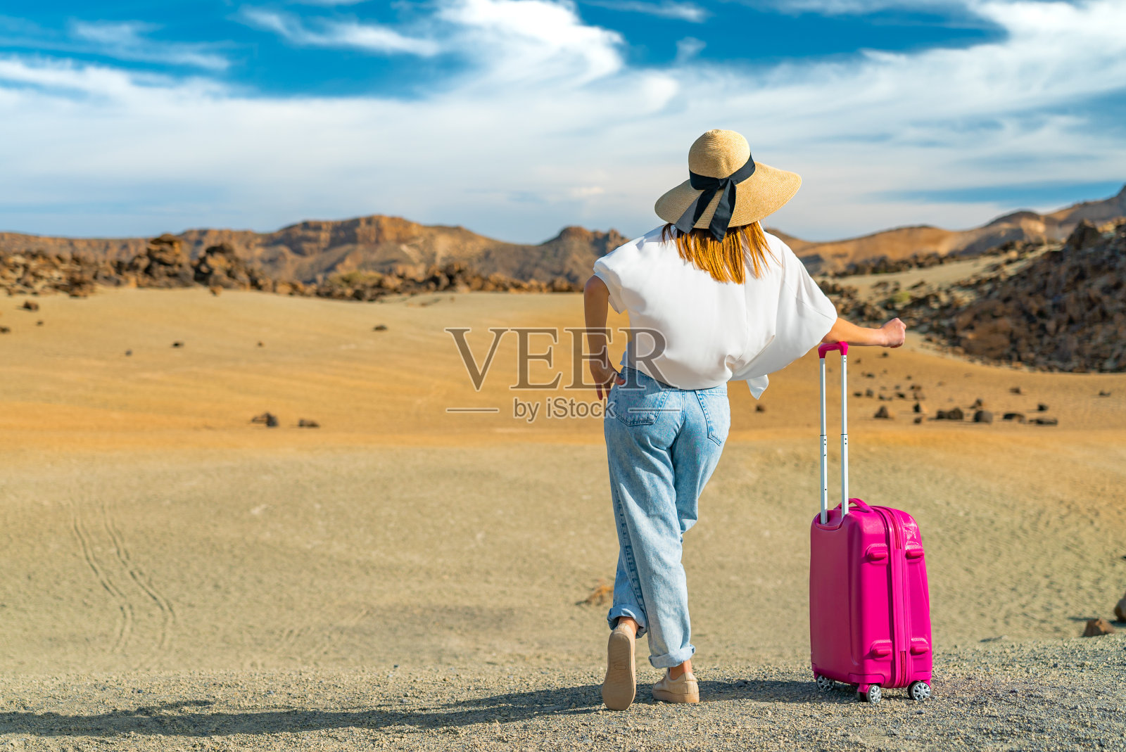 女孩站在一个白色的衬衫和帽子和一个粉红色的手提箱在火山沙风景。泰德火山,特内里费,西班牙。照片摄影图片