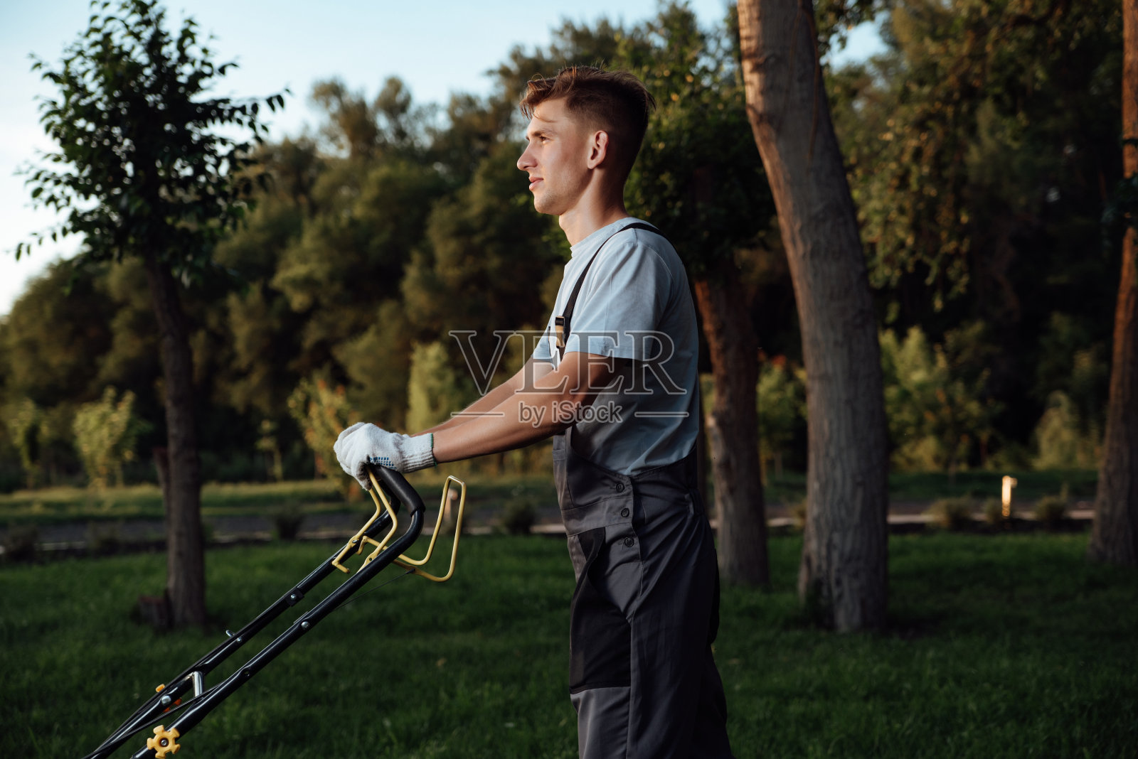 一个穿着工装裤的年轻男园丁正在使用割草机。照片摄影图片