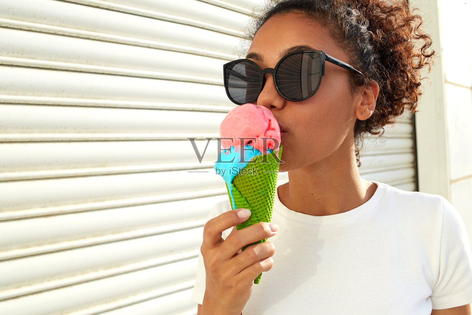 在一个阳光明媚的日子里,一个年轻美丽的非洲裔美国女孩身穿白色t恤和浅色牛仔裤,靠着一面墙吃着冰淇淋。照片摄影图片