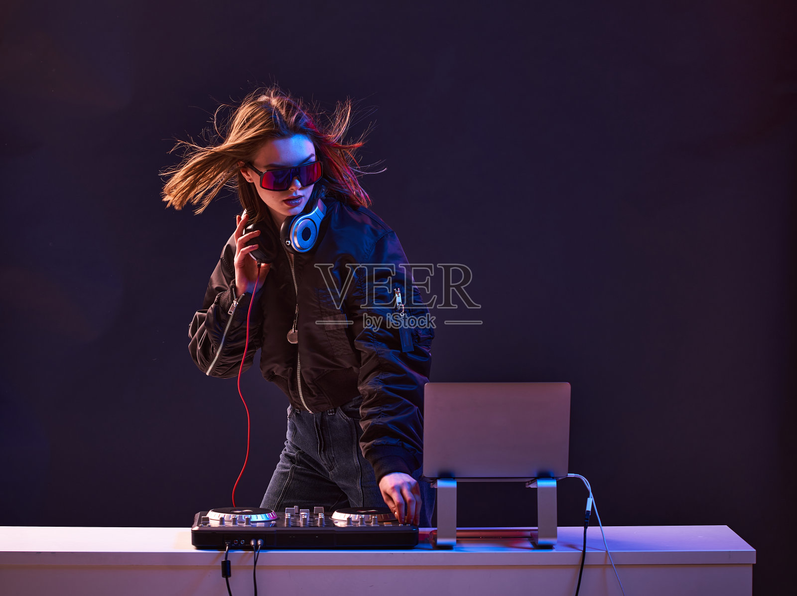 时尚的DJ女孩戴着耳机和眼镜,穿着短夹克,将派对上的音乐和霓虹灯混合在一起。照片摄影图片