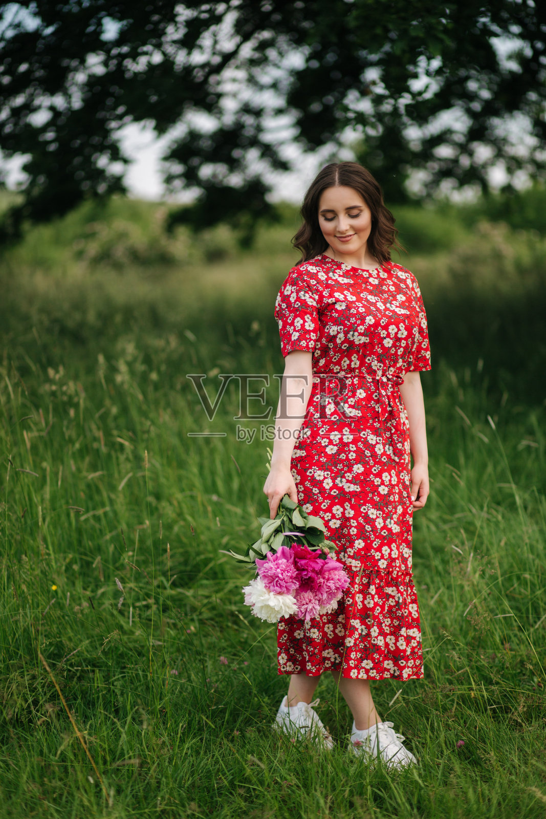 有魅力的年轻女子穿着红色的裙子,带着美丽的花朵走在大树前的田野里照片摄影图片