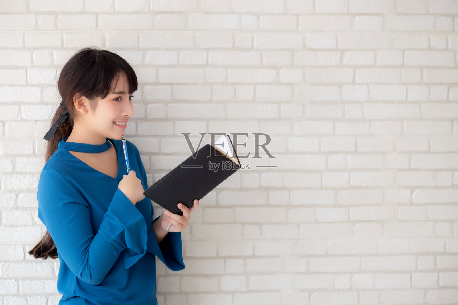 美丽的亚洲女人微笑着站在家里水泥白色的背景上思考和写笔记本,女孩作业书上,教育和生活理念。照片摄影图片