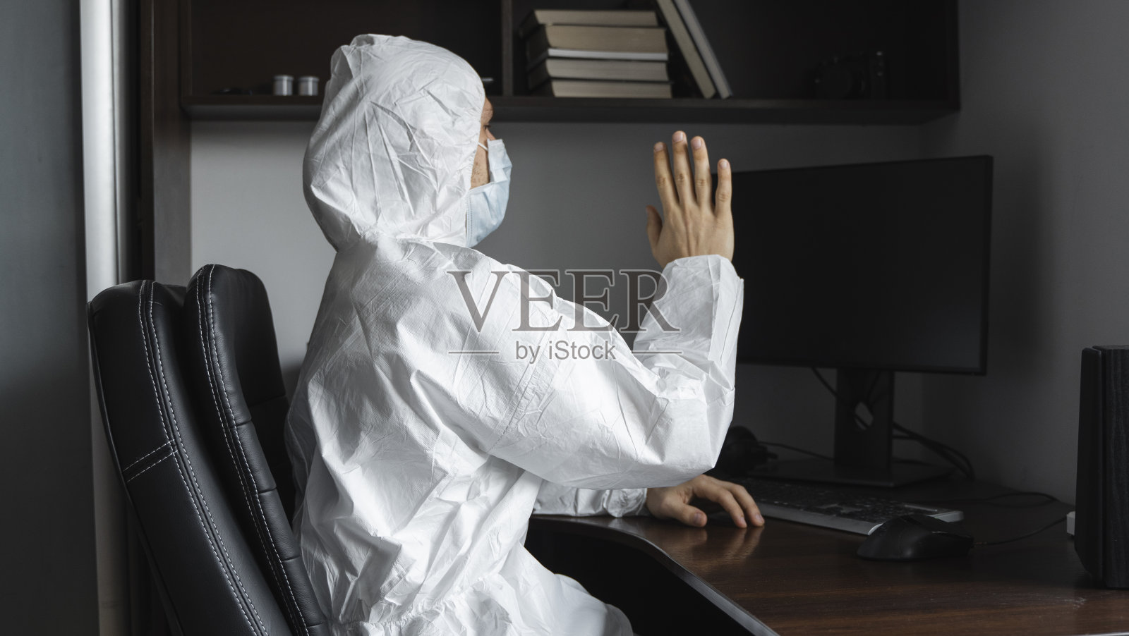 在隔离期间,戴着医用口罩和防护服、手套的人坐在家里,用电脑在桌子旁工作,向他的商业伙伴或朋友问好。冠状病毒大流行中的远程工作。照片摄影图片