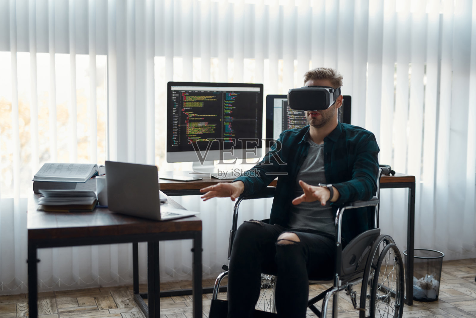 虚拟现实在起作用。年轻男性网络开发人员坐在轮椅上,戴着虚拟现实眼镜,在他的现代办公室工作。高科技照片摄影图片