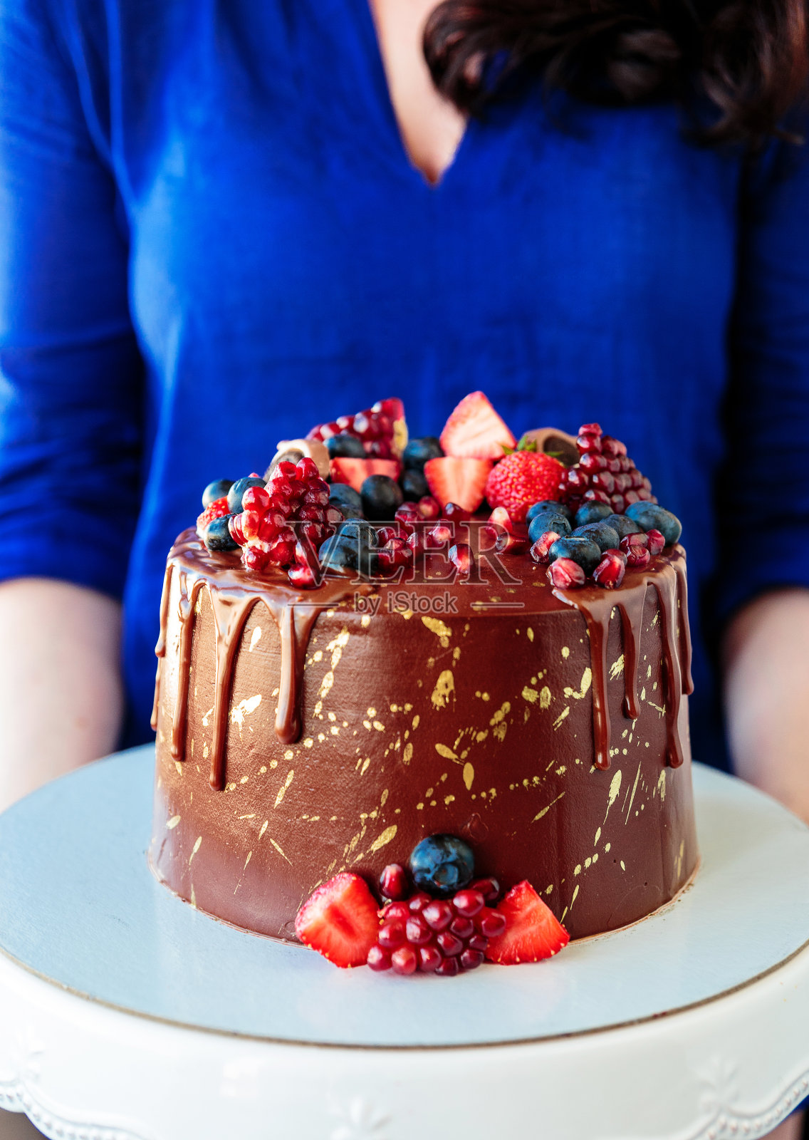 一位身穿蓝色衬衫的女士手里拿着一个托盘,托盘上有一个美味的巧克力蛋糕,上面装饰着草莓、蓝莓和石榴。照片摄影图片