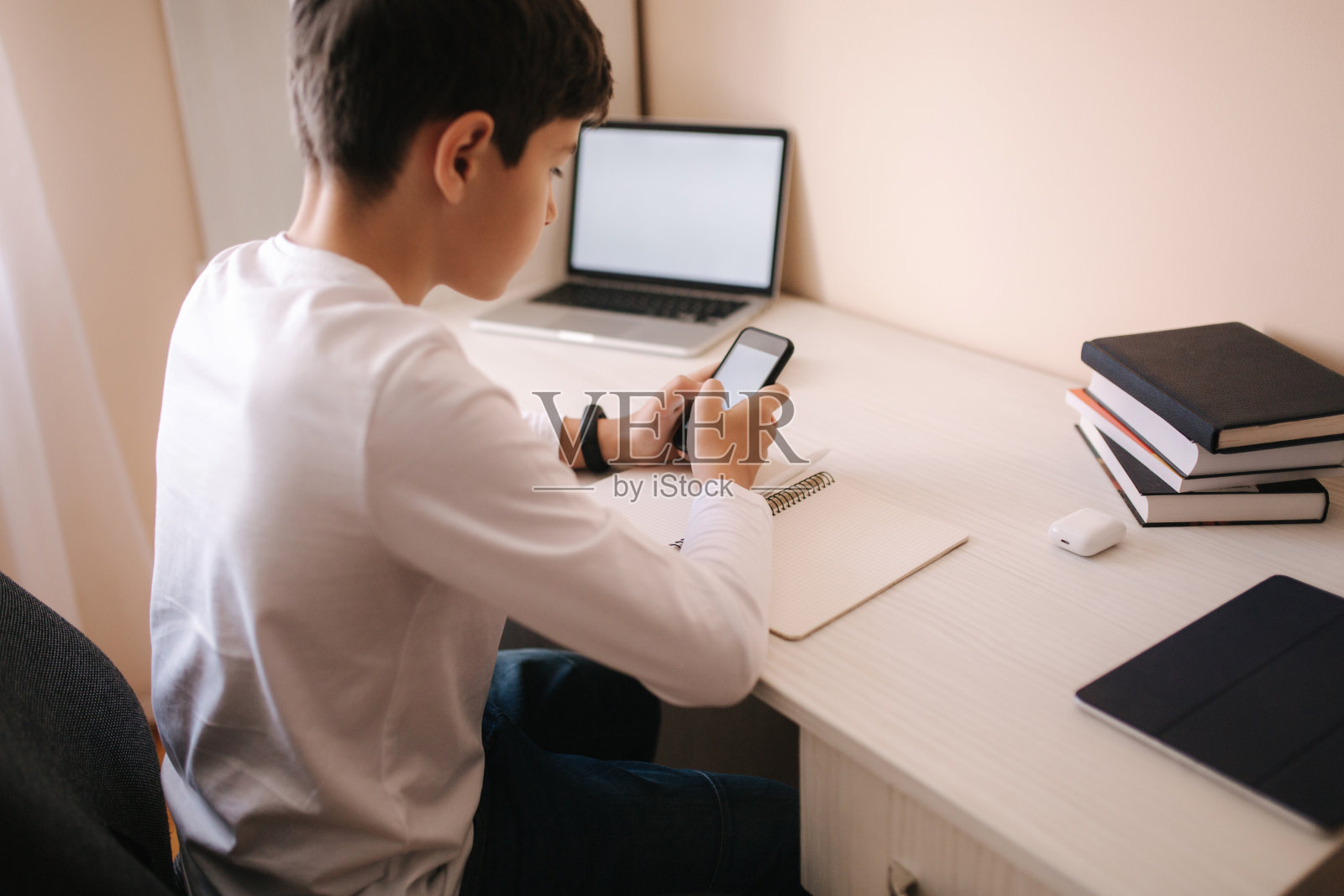 一个男生在他房间的书桌前学习。男孩用笔记本电脑和智能手机在笔记本上写字。书和写字板放在桌子上。在隔离期间在家学习照片摄影图片