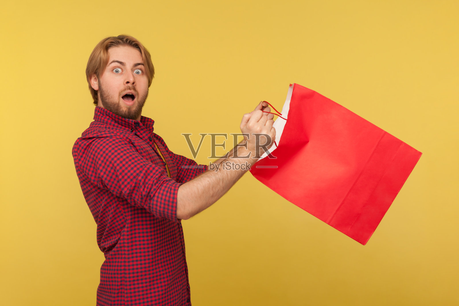 侧视图惊讶的购物男子在格子衬衫拿着购物袋,看着相机与大眼睛照片摄影图片
