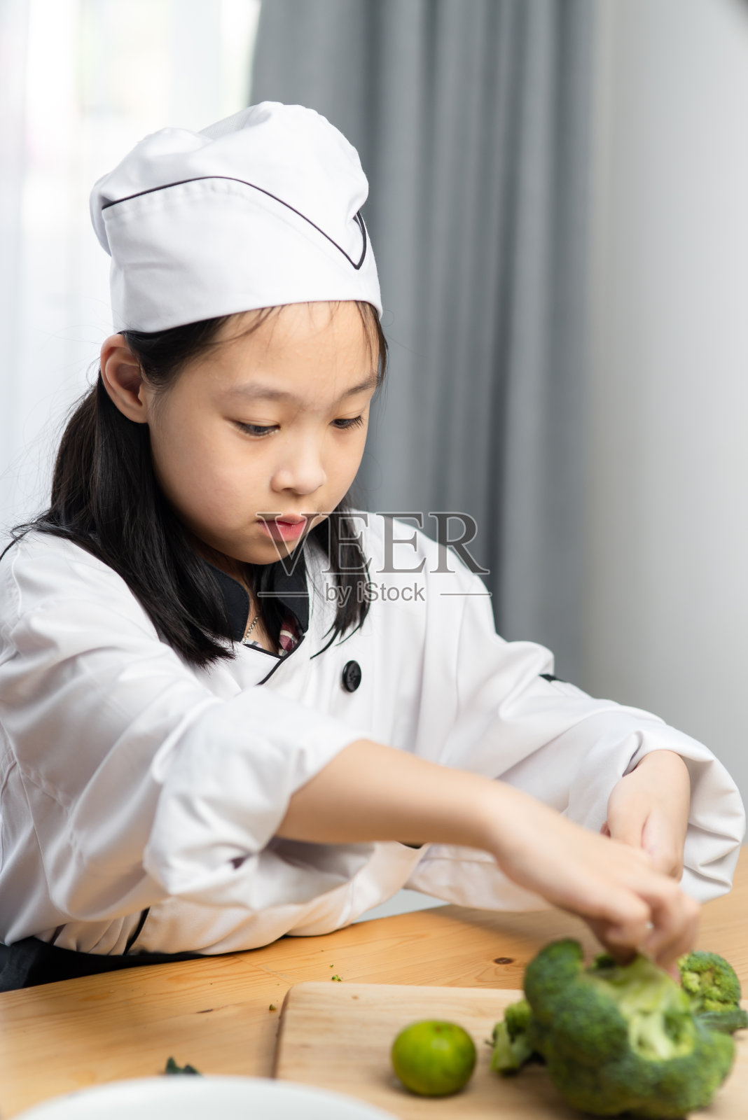 快乐的亚洲女孩穿着厨师服在家切西兰花。照片摄影图片