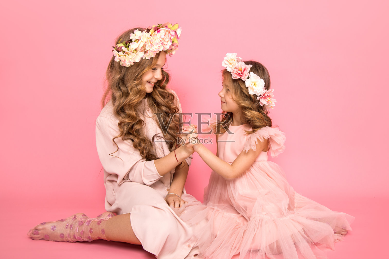 照片中的两个美丽的女孩坐在粉红色的背景看着对方手牵手。姐姐和妹妹之间的关系。家庭。爱。友谊。花环的花照片摄影图片