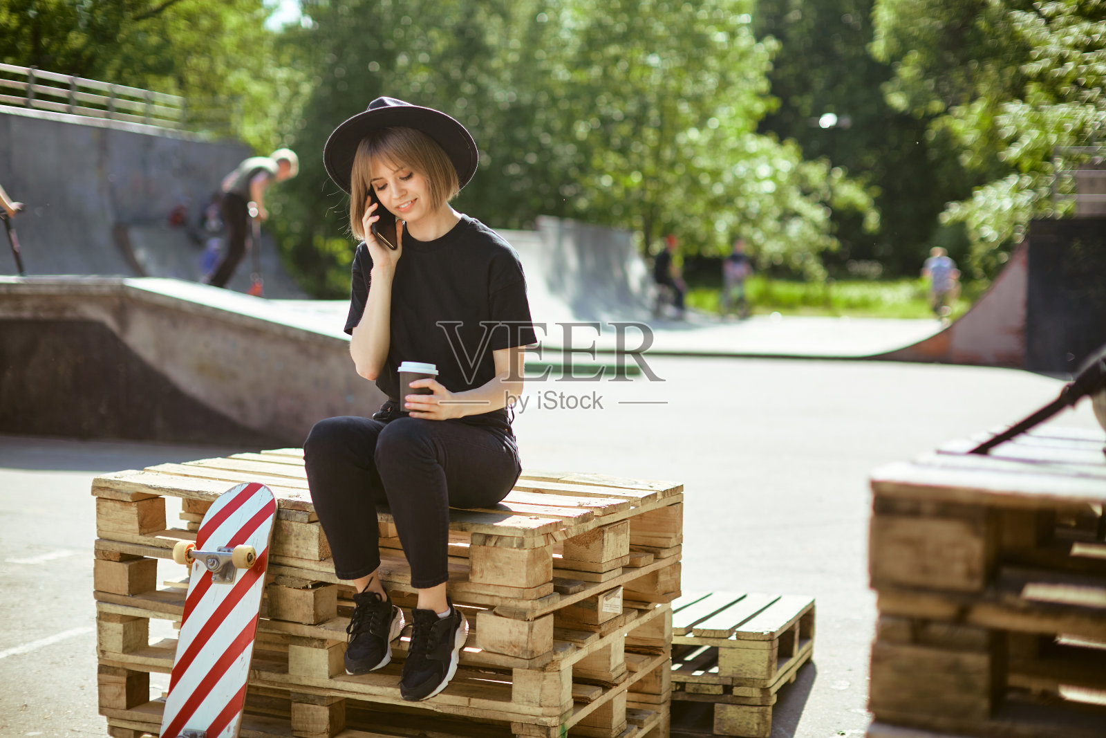 戴着帽子的年轻女孩博主坐在公园的木托盘上,用滑板和杯咖啡进行电话交流照片摄影图片