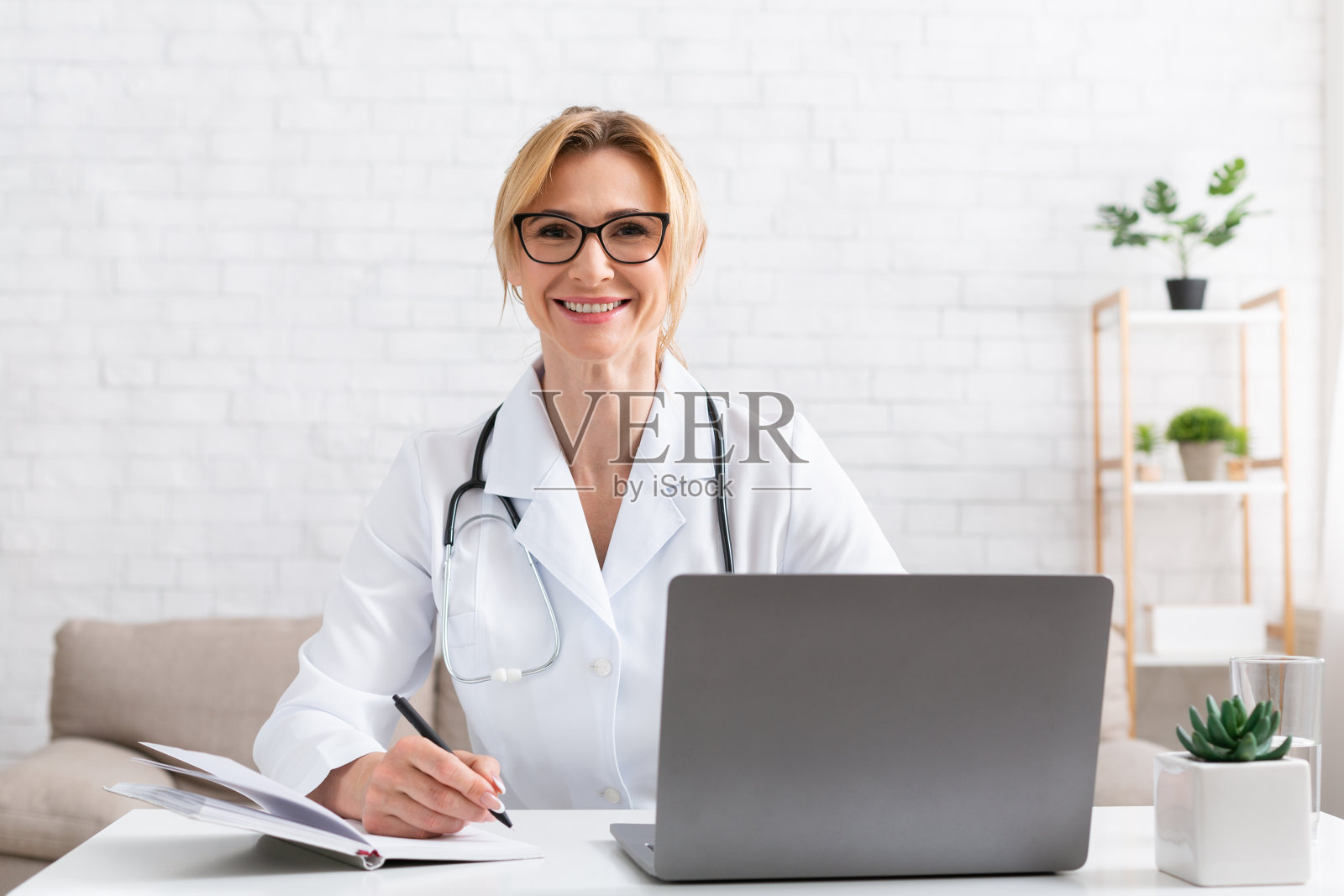 在线治疗师。微笑的女医生在工作场所与笔记本电脑和记事本照片摄影图片