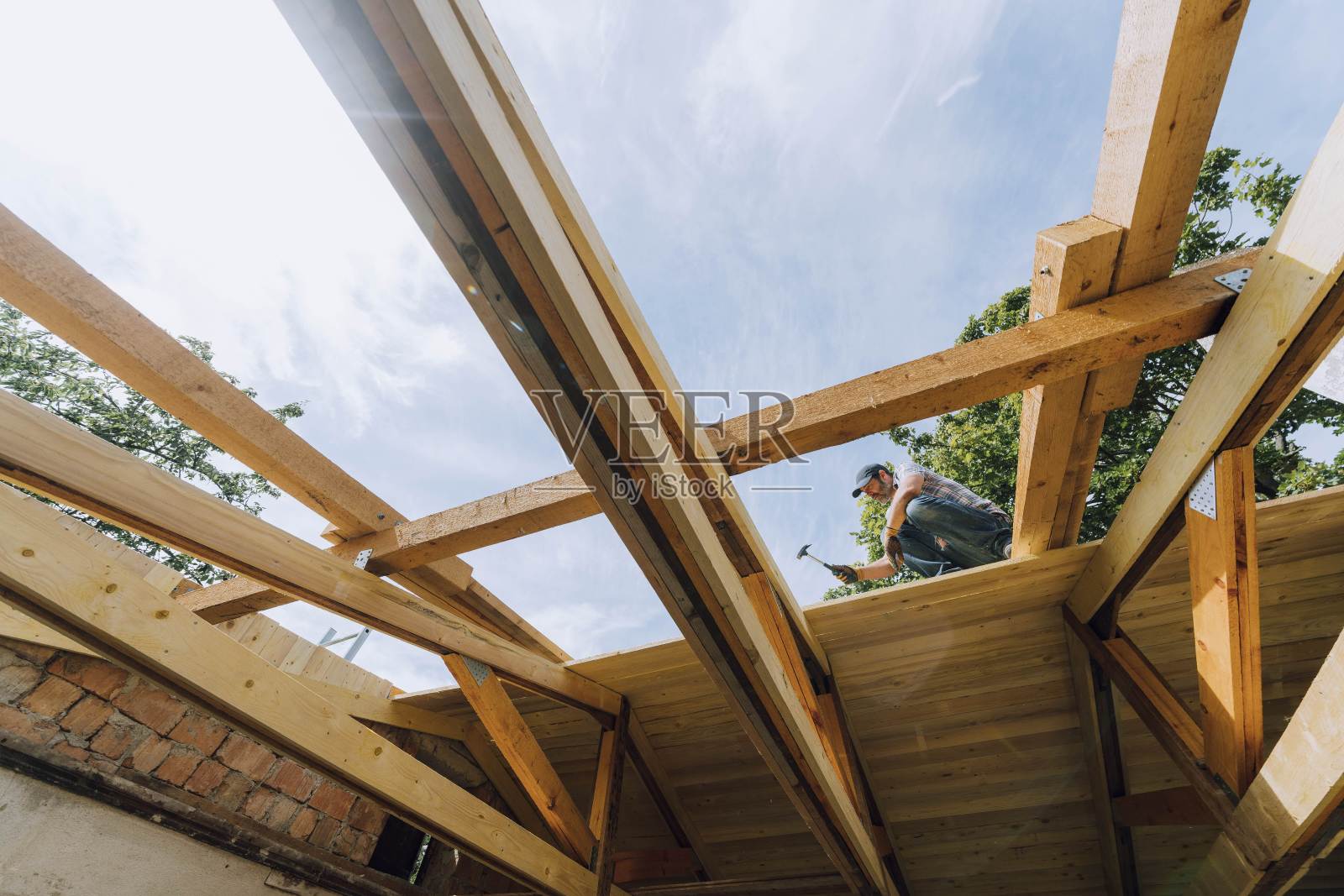 现场未完工住宅结构的木梁上,男木匠在用锤子敲击时蹲下照片摄影图片