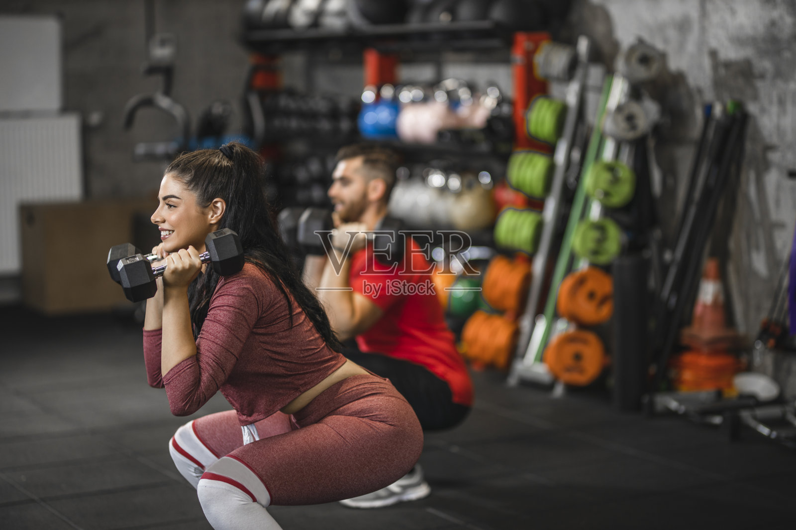 喜欢运动的年轻人在健身房用哑铃和微笑锻炼,喜欢运动的年轻人做负重蹲坐照片摄影图片