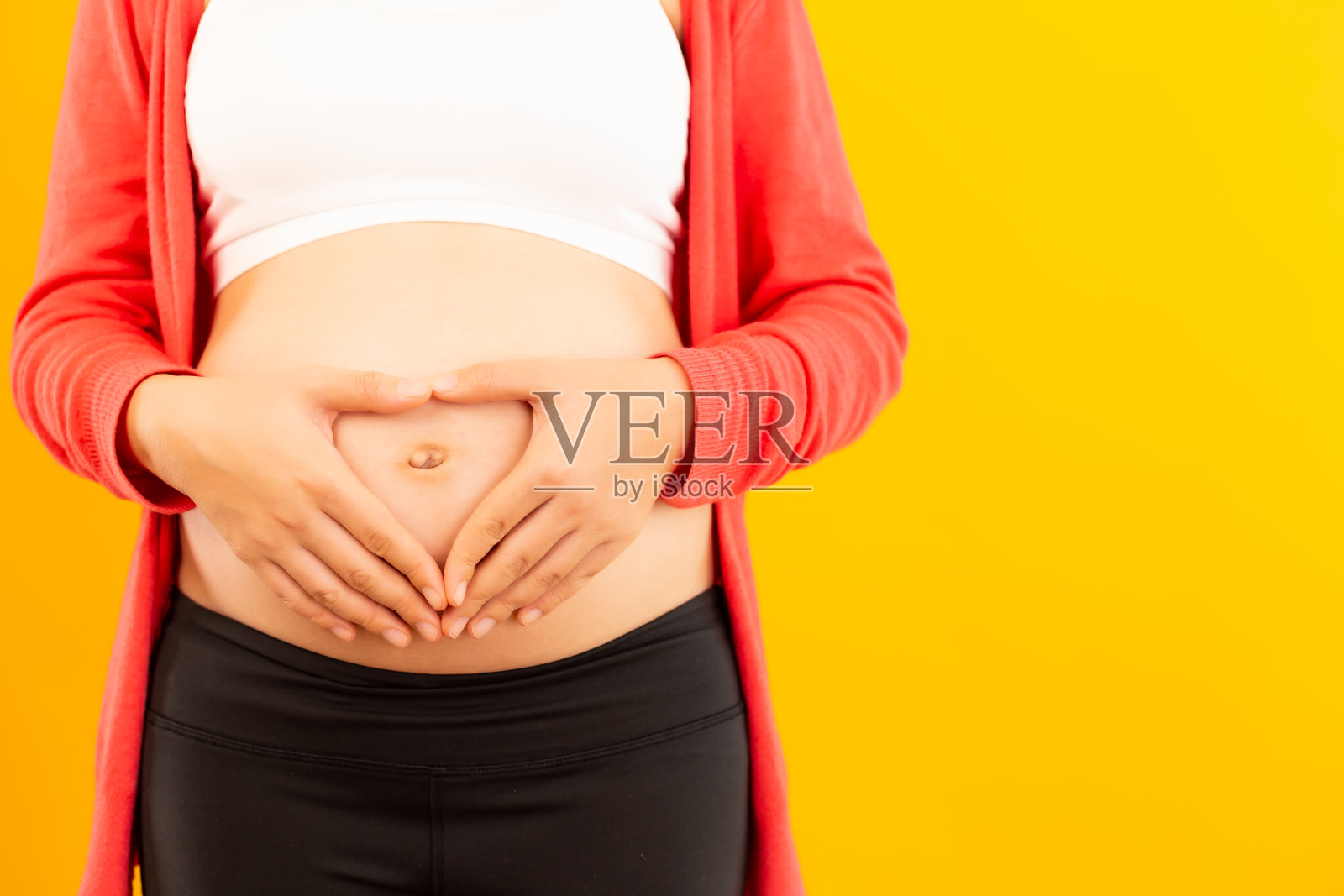 快乐的年轻孕妇用手比心,放在腹部上,表达对宝宝的爱。产前、怀孕、母爱、期待概念,黄色背景,留白照片摄影图片