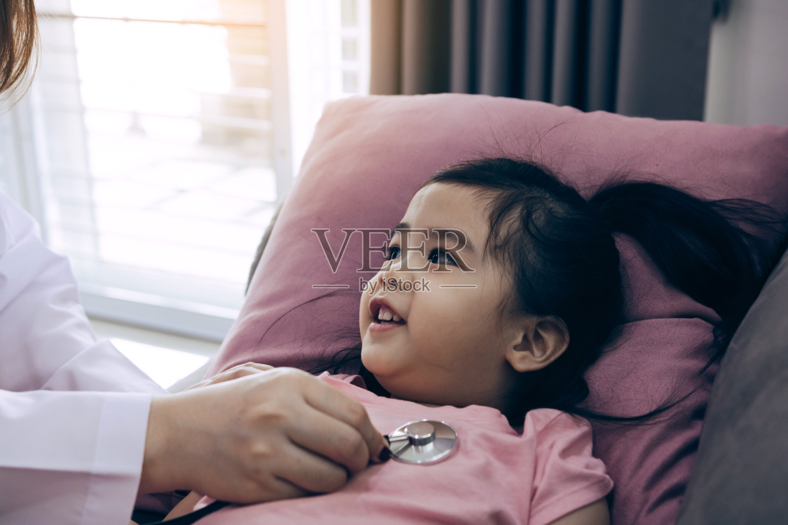 一个亚洲小女孩,一个女人睡在沙发上的枕头上,一个女医生在家里用听诊器检查心跳。照片摄影图片