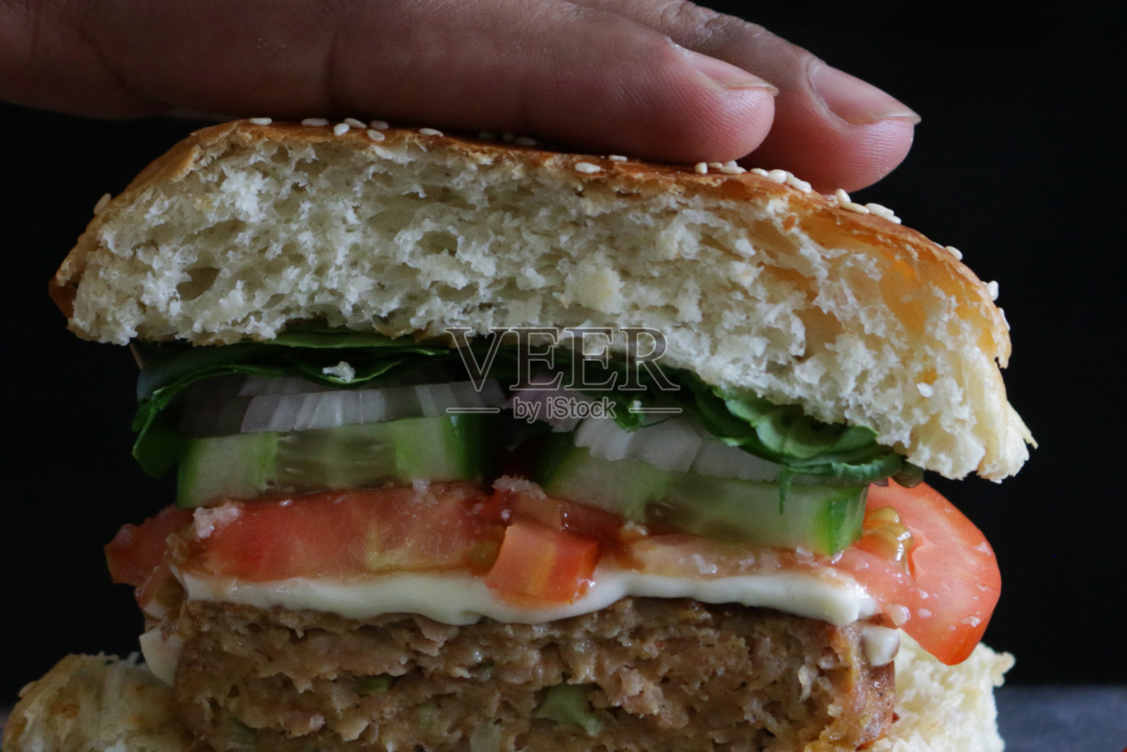 特写镜头:一个不认识的人拿起自制的汉堡,夹着芝麻、融化的奶酪、切成薄片的番茄、黄瓜、洋葱圈和莴苣的面包,牛肉汉堡切成两半放在石板铺的垫子上,黑色背景照片摄影图片