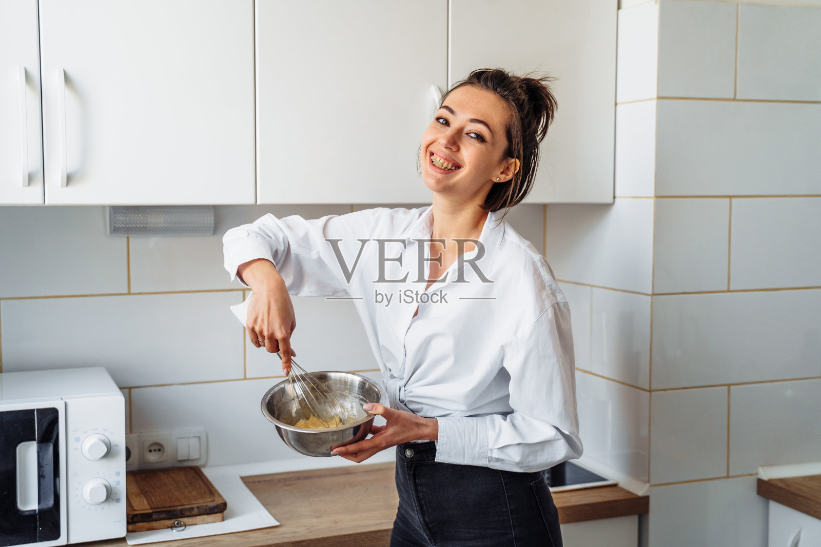 美丽,迷人,微笑的年轻女子在一个白色,明亮的厨房里,用搅拌器在钢盘子里揉香蕉煎饼的面团。黑白照片没有明亮的口音照片摄影图片