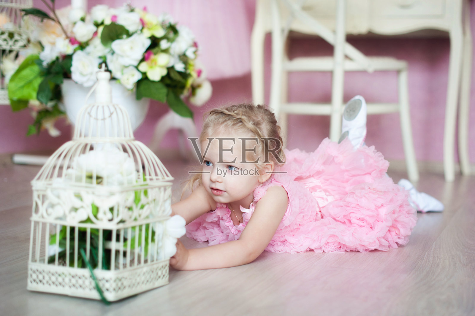 一个穿着粉红色裙子的小芭蕾舞女演员躺在舞蹈教室的地板上,审视着一个插满鲜花的笼子。照片摄影图片