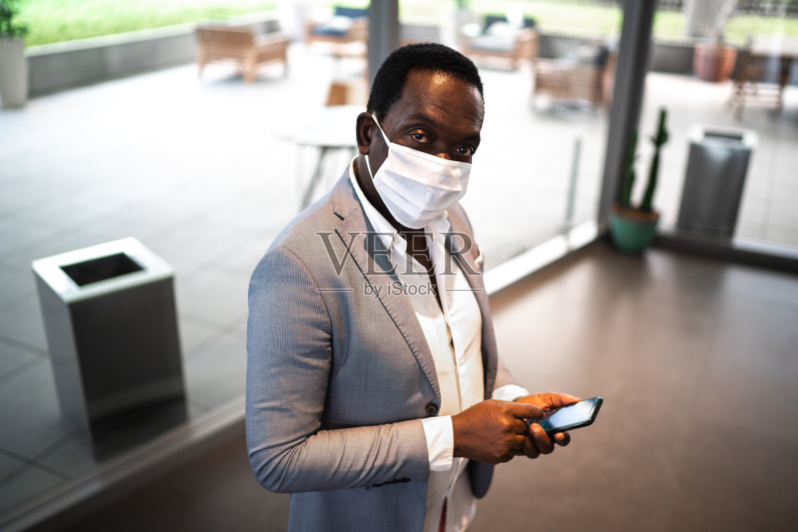 在办公室大厅里用手机等电梯的成熟男子的肖像——带着面具照片摄影图片
