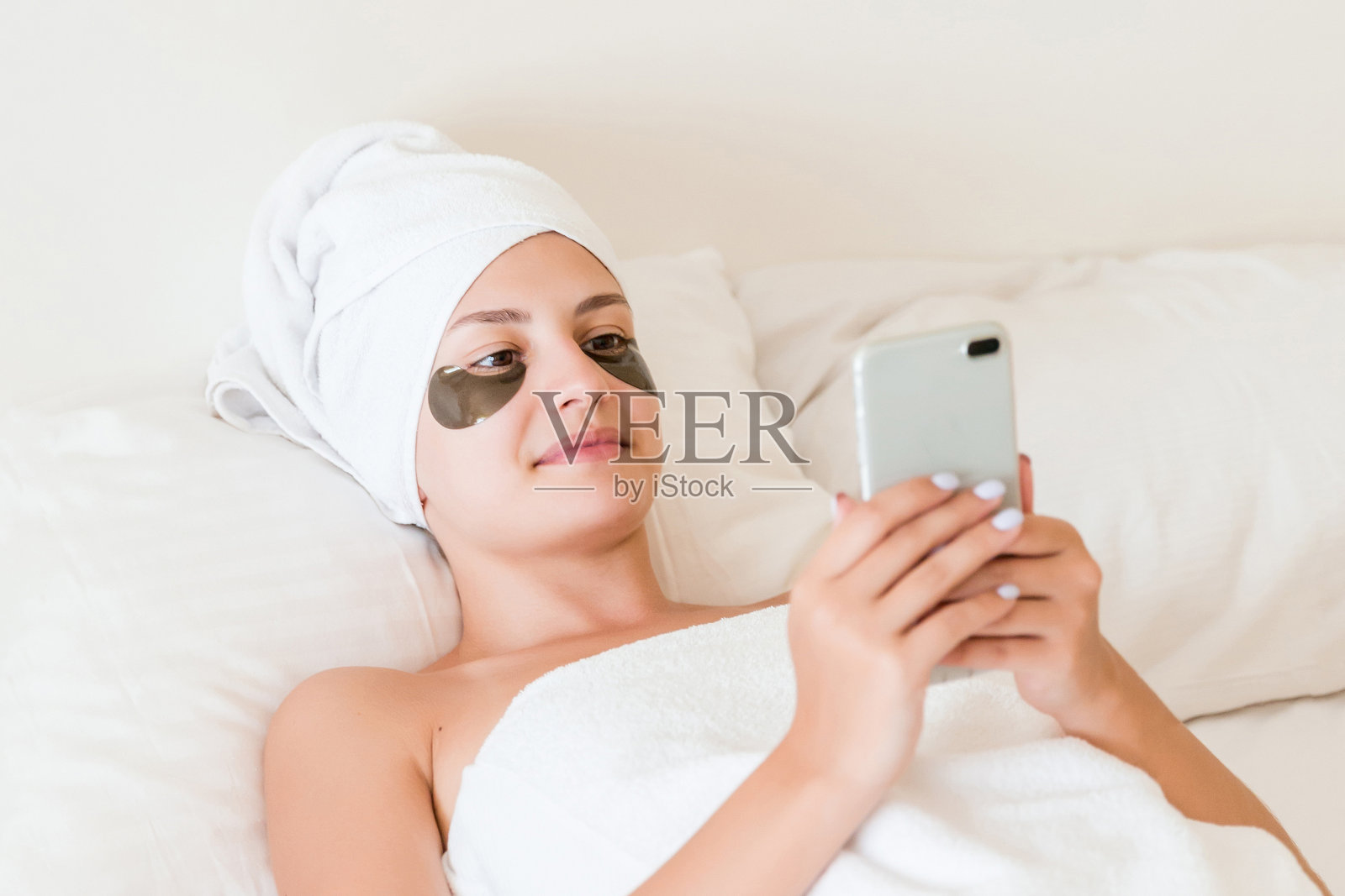 年轻女子沐浴后裹着毛巾和眼罩在床上放松和使用她的手机。女孩正在读信息。完美清新的肌肤理念照片摄影图片