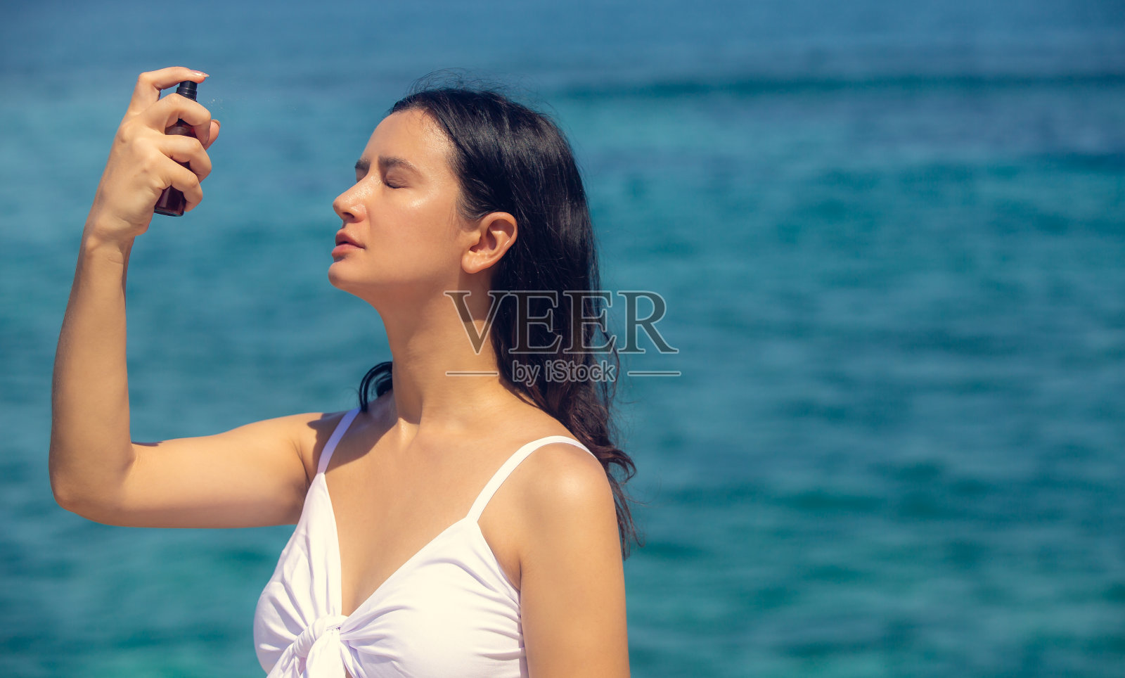 年轻漂亮的女人在海滩上喷洒热水或面部喷雾在她的脸上。夏季皮肤护理照片摄影图片