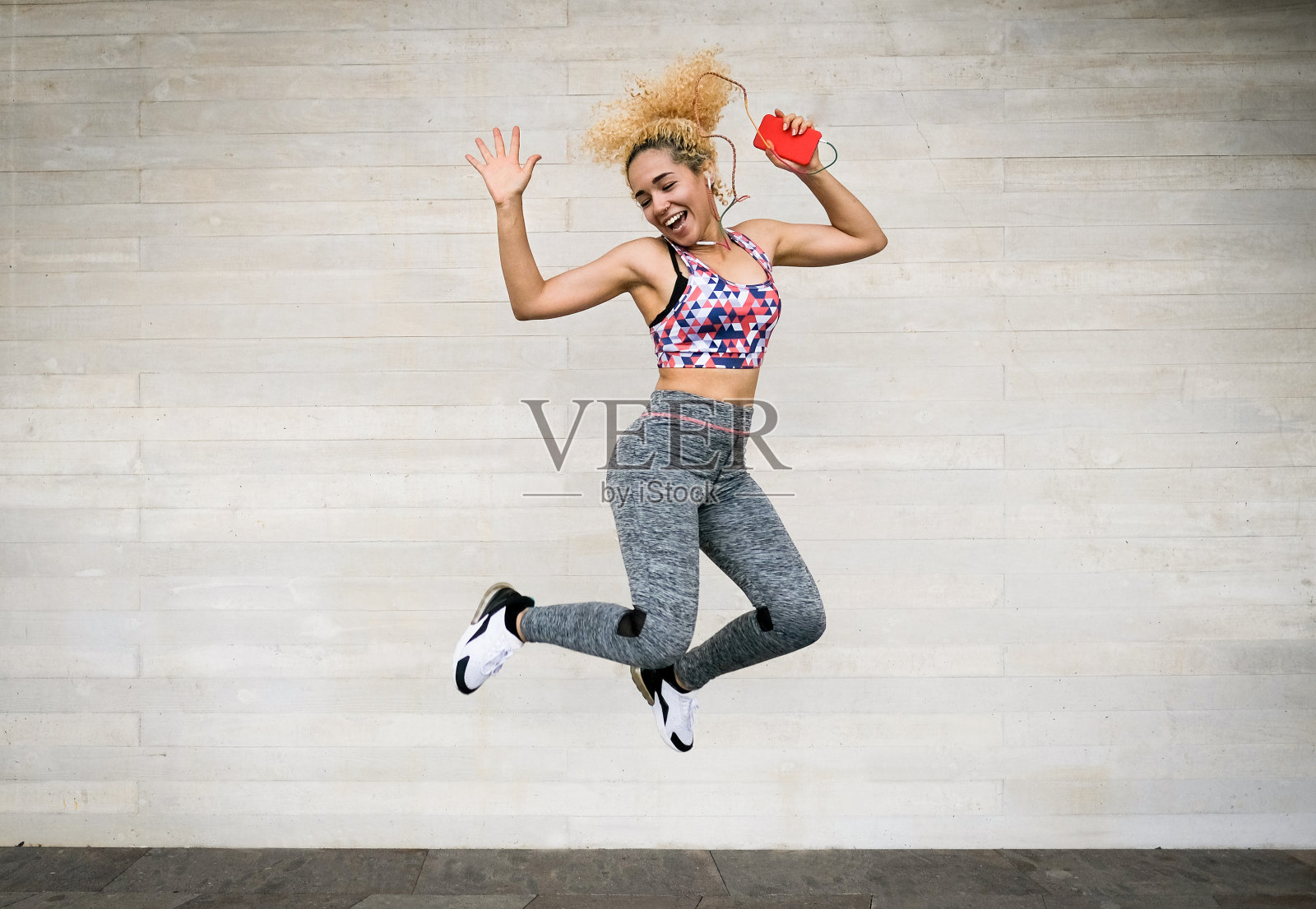 年轻的多种族女子听音乐从播放列表和跳跃-快乐的运动女人与耳机跳舞,在她的手中拿着智能手机照片摄影图片