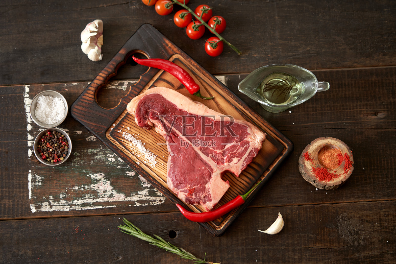 新鲜、陈年的t骨牛排放在砧板上,配上两个红辣椒、橄榄油、干番茄和红辣椒粉、盐和胡椒、一根番茄和大蒜,放在木质的深色背景上,放置文字。木炭烹饪照片摄影图片