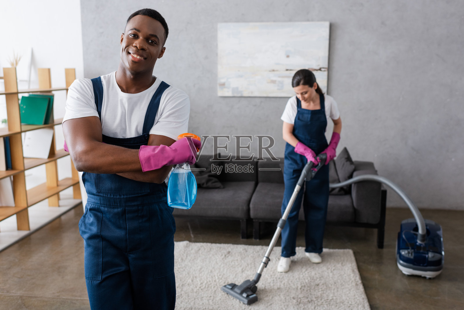 非裔美国清洁工拿着洗涤剂,对着镜头微笑,旁边的同事正在清洗地毯照片摄影图片