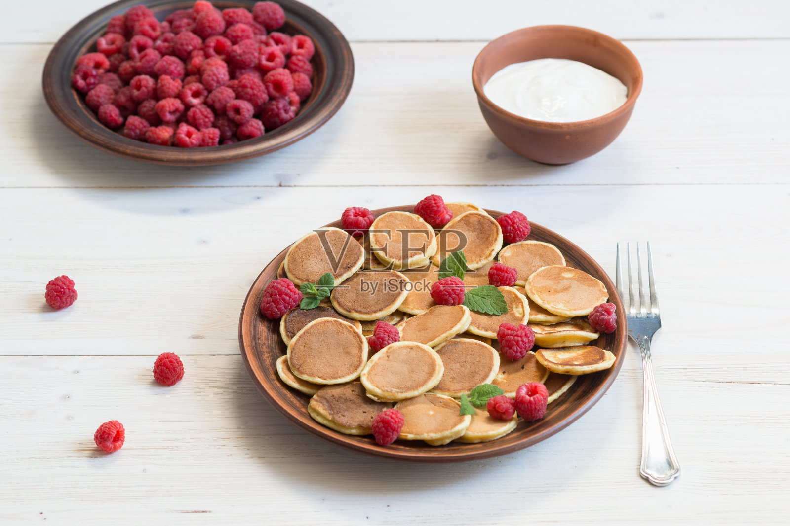 白色木桌上的棕色盘子里盛着麦片煎饼、酸奶和成熟新鲜的覆盆子。美味的早餐照片摄影图片