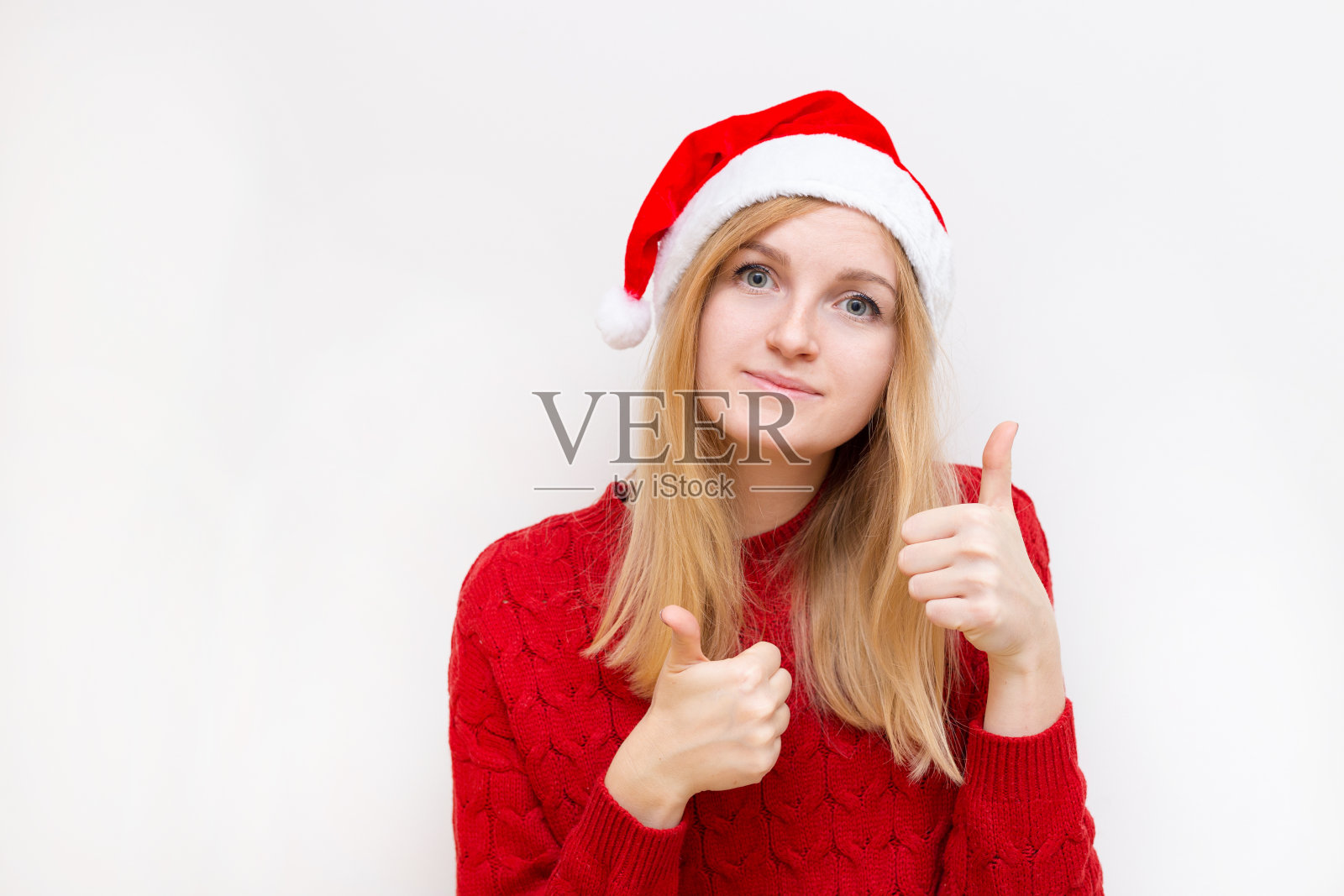 戴着圣诞帽的快乐女人竖起大拇指。女孩看着相机和微笑孤立在白色的背景。照片摄影图片