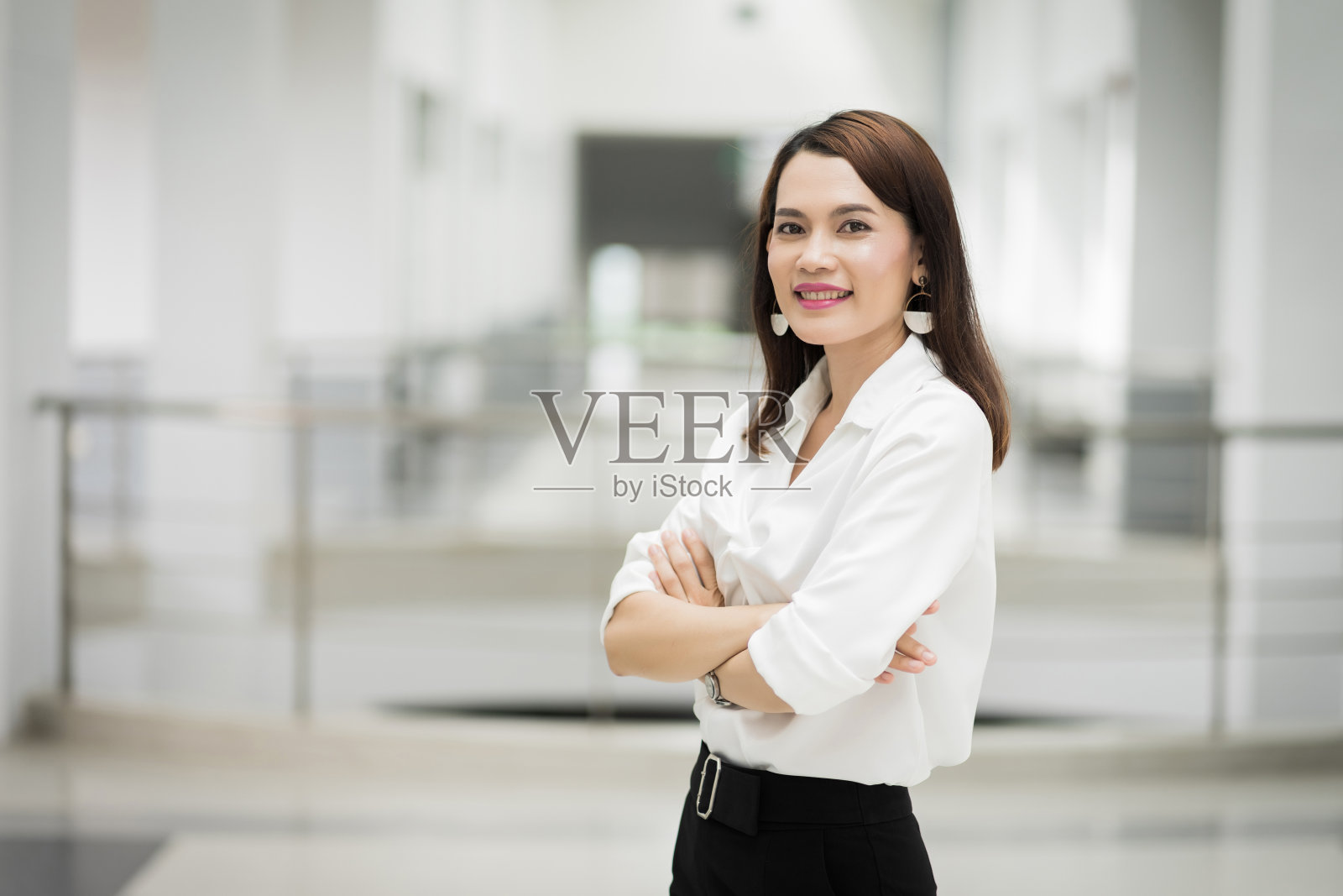 一个快乐的中年女商人穿着商务套装,双臂交叉站在公司大楼里。现代商业女性在有复印空间的办公室。商业女性获得stoc照片摄影图片