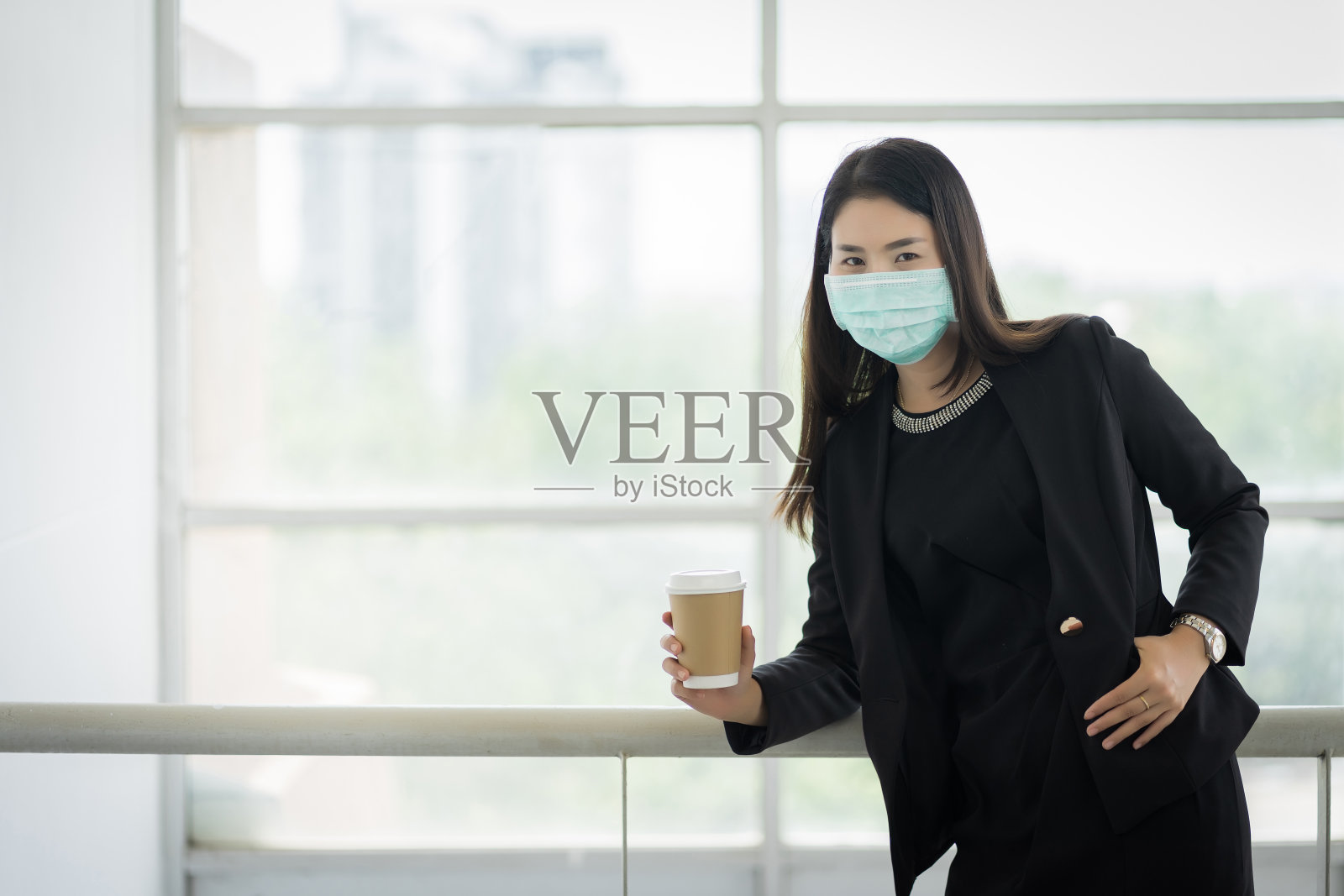 一名穿着西装、戴着口罩的女商人在公司大楼里拿着早上的咖啡杯准备工作。一名中年妇女戴着口罩预防COVID-19大流行。库存图片照片摄影图片