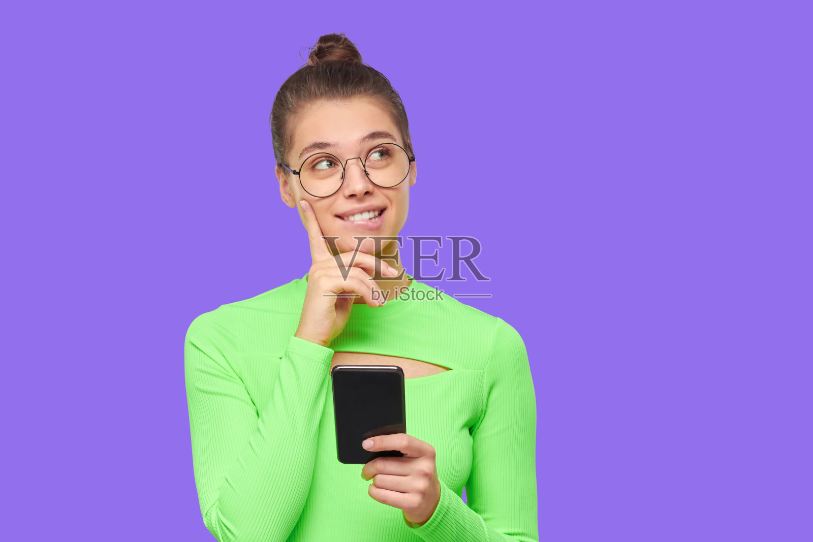 梦想中的女孩,脖子上戴着眼镜和耳机,微笑着看向别处,手里拿着手机,孤立在紫色的背景下照片摄影图片