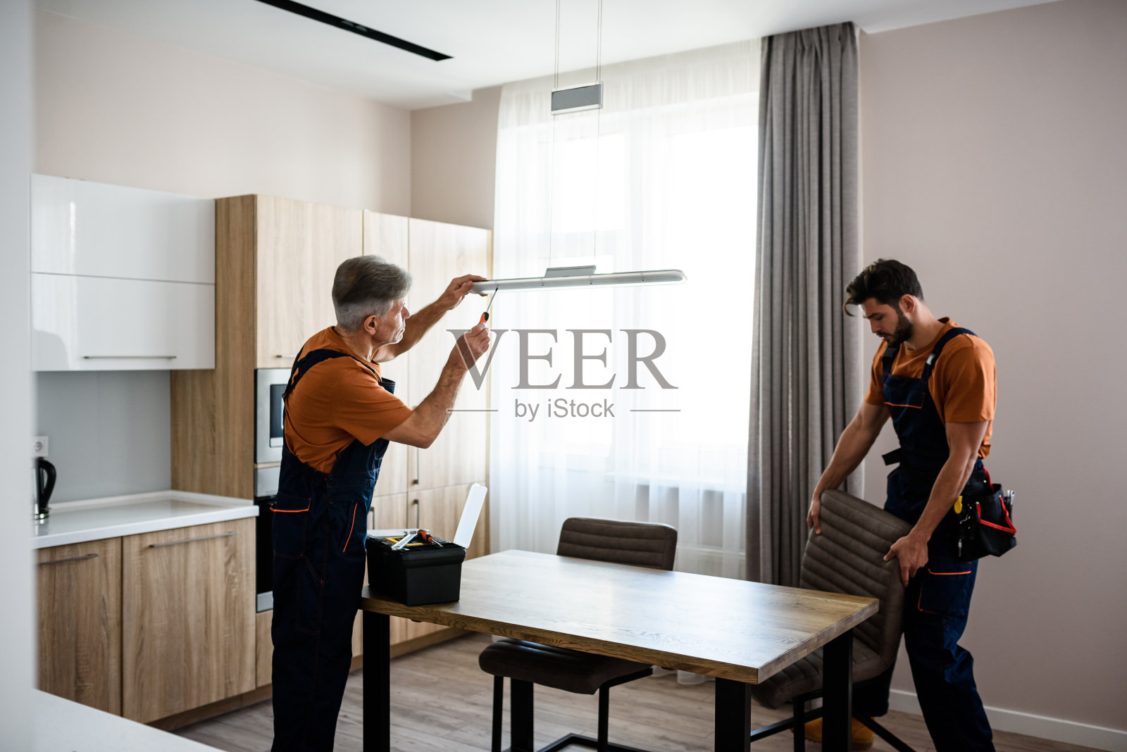 我们让它变得更好。两个杂工,工人制服固定,安装灯和设备在厨房,使用螺丝刀在室内。维修和装配服务理念照片摄影图片