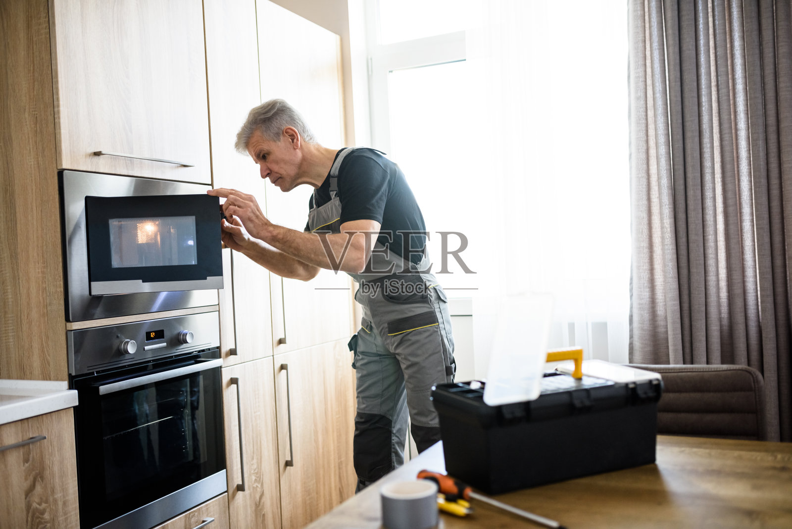 穿着制服的老修理工在厨房里用手电筒检查坏掉的微波炉。维修服务理念照片摄影图片