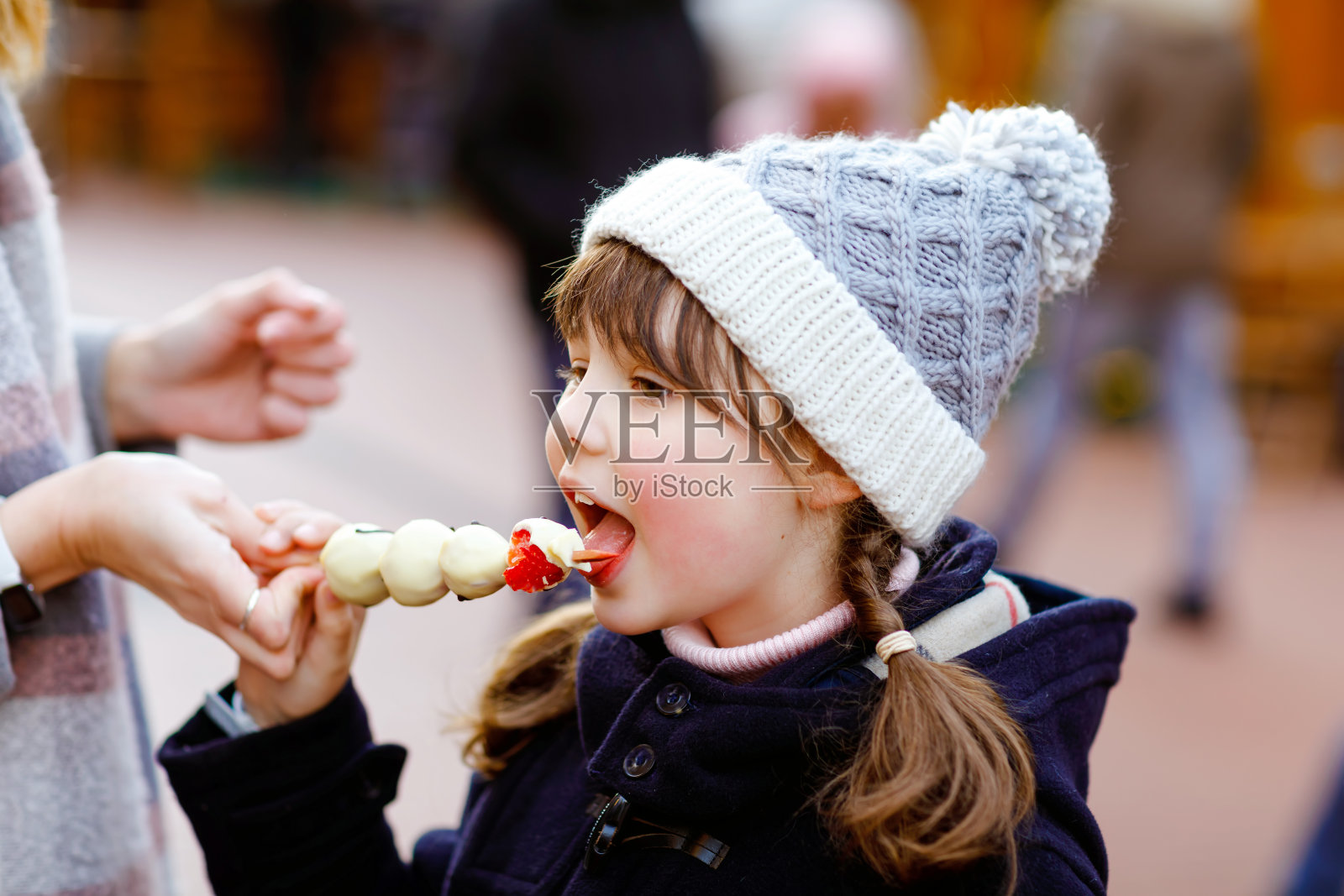 德国传统圣诞市场上,可爱的小女孩正在吃白巧克力串草莓。快乐的孩子在传统家庭市场在德国雪天。照片摄影图片
