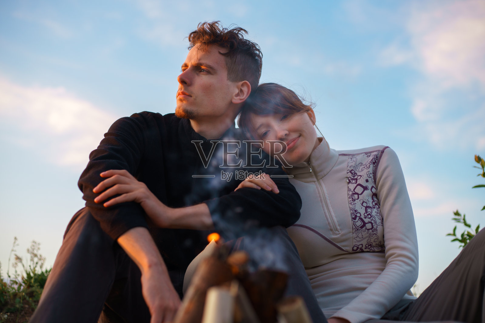 一对年轻的夫妇坐在一根倒下来的木头上,互相拥抱着,看着火烧起来照片摄影图片