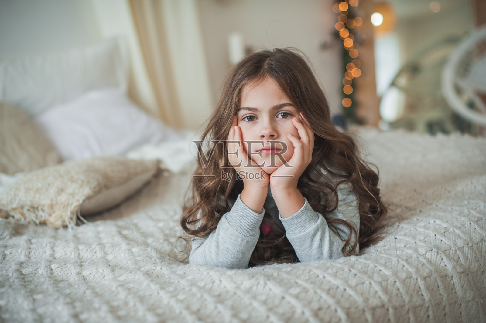 漂亮的小女孩穿着灰色睡衣躺在床上,留着长长的黑色卷发,用手撑着脸,看着镜头。软的焦点。照片摄影图片