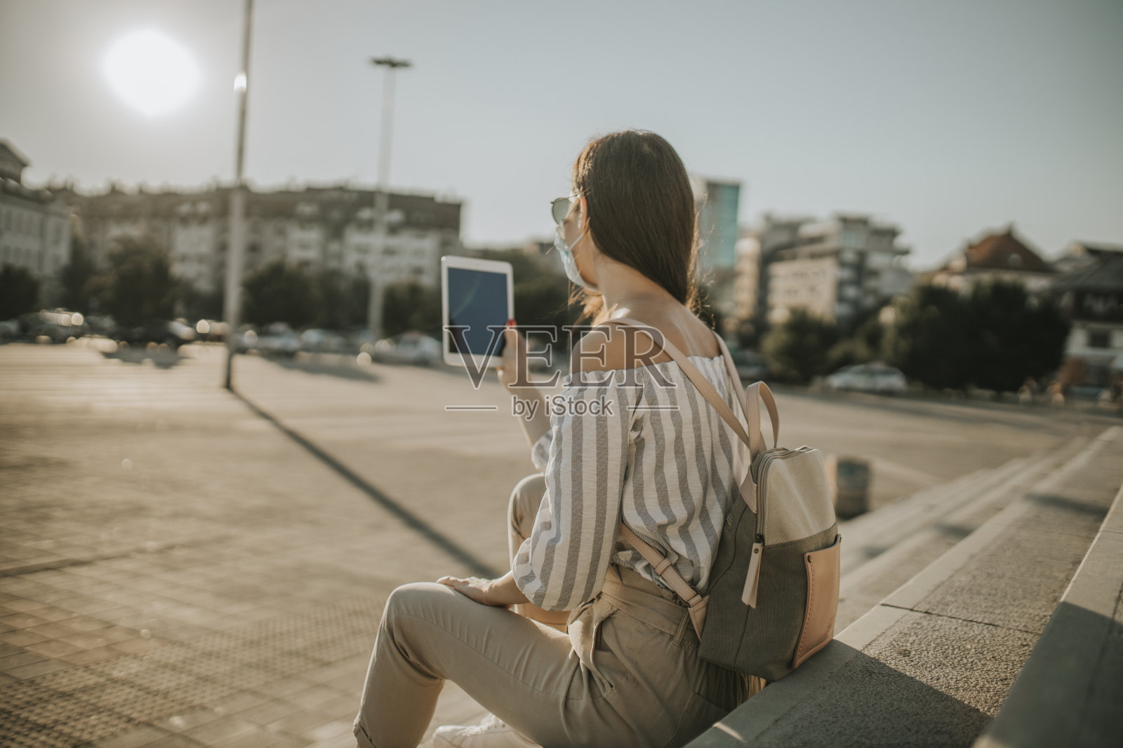 在冠状病毒大流行期间,戴医用口罩的妇女在城市使用电子平板电脑,坐在楼梯上照片摄影图片