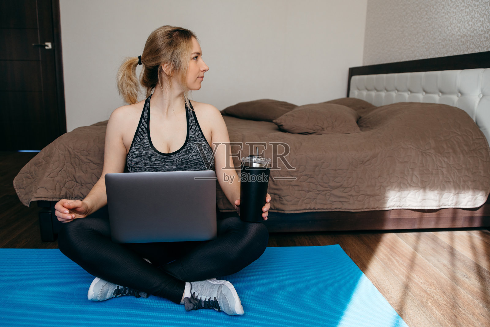 一位穿着运动服的运动女性正坐在地板上,手里拿着哑铃、蛋白奶昔或一瓶水,正在起居室里使用笔记本电脑。运动和娱乐的概念。照片摄影图片