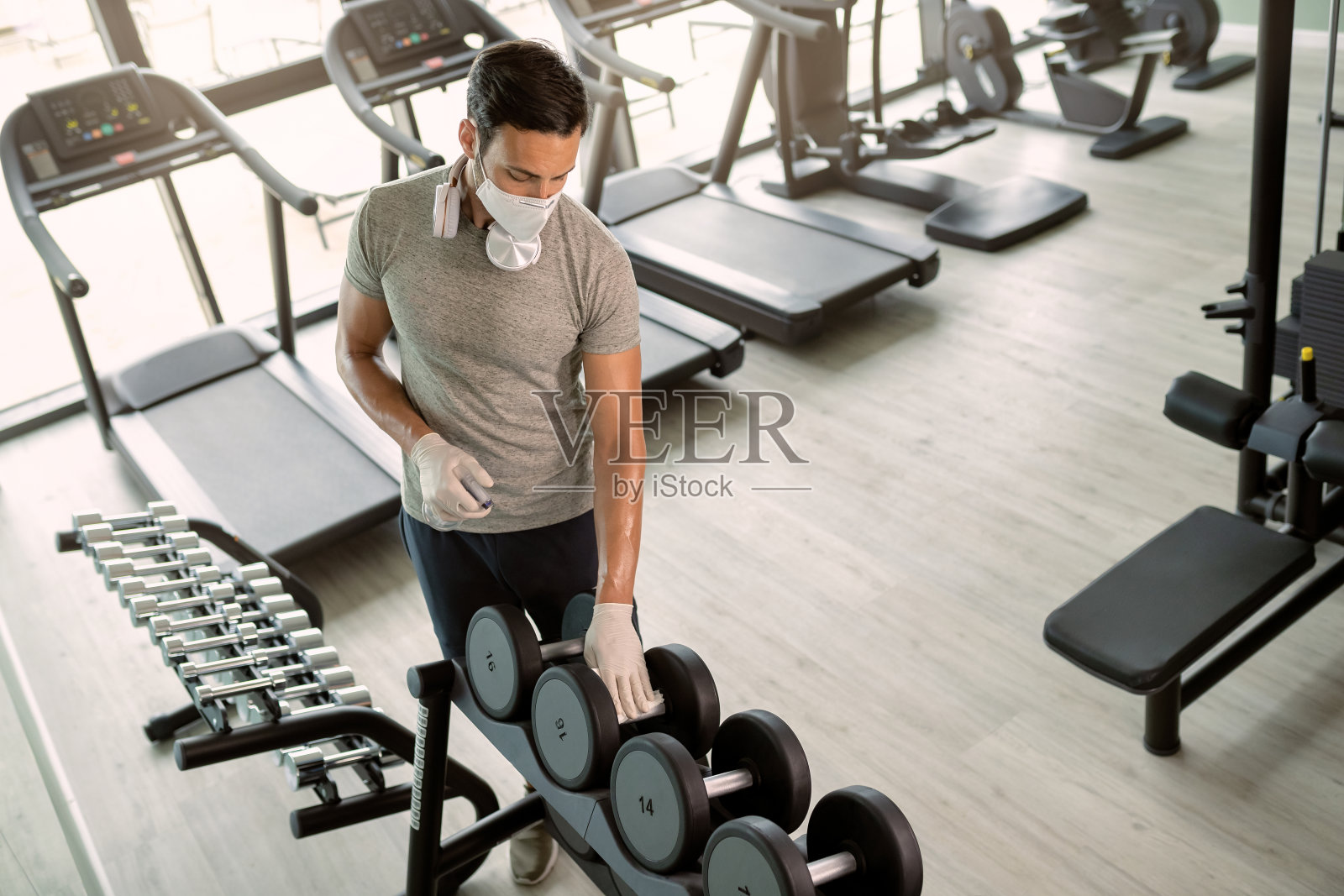 运动员戴防护面罩,在健身俱乐部用消毒剂清洁哑铃照片摄影图片