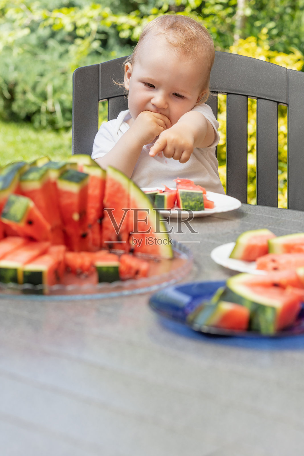 可爱的小男孩正在吃东西,他的手指着桌子上的一个西瓜。照片摄影图片