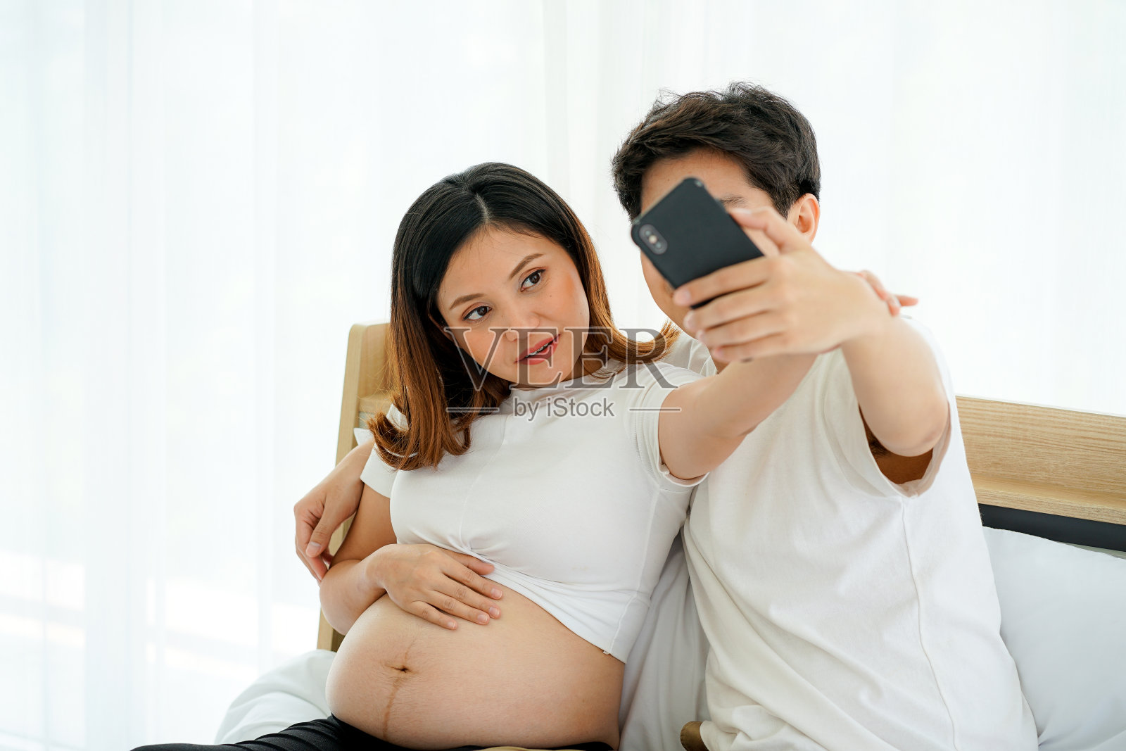 年轻的亚洲孕妇和丈夫在房子里的白色床上用手机自拍照片摄影图片
