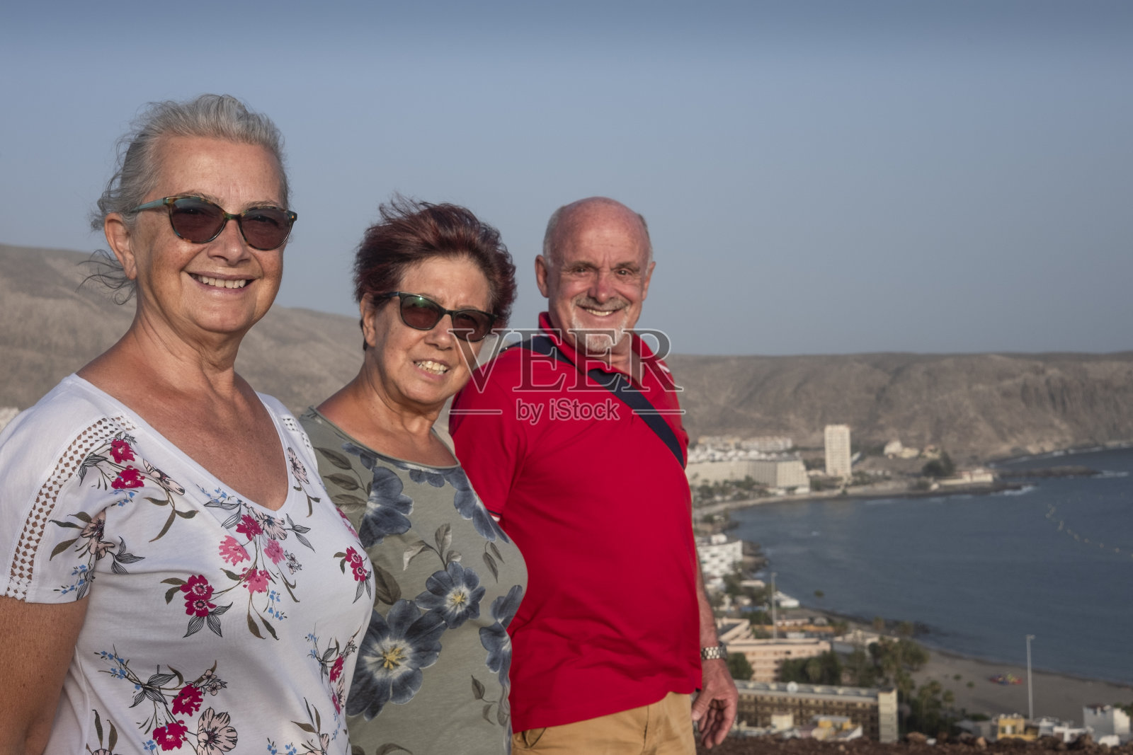 一组三个微笑的老年人站在小山对面的海滩上看着照相机和海上活跃的老年人和假日概念照片摄影图片